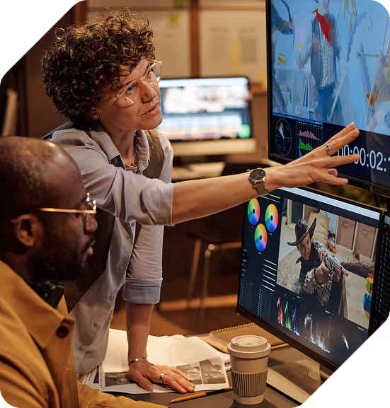 Two people collaborating in a video editing studio, with one pointing at a monitor showing edited video content and color grading tools.
