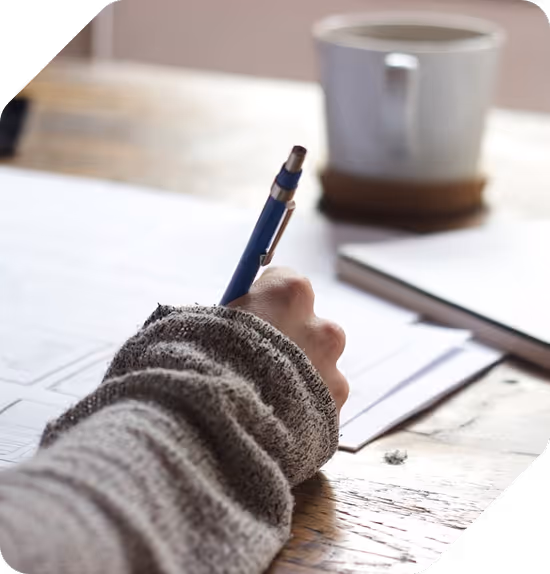 Hand in a gray sweater holding a blue pen, writing on paper on a wooden table with a blurred cup in the background.