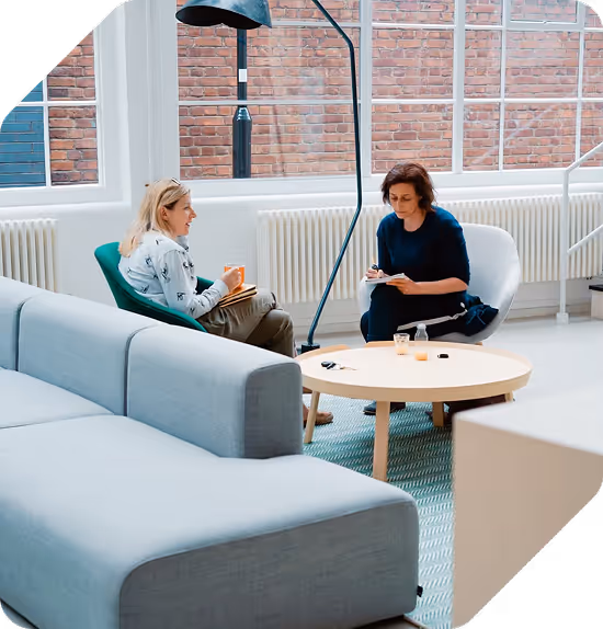 Two women sitting and talking in a bright modern living room with large windows and a gray sofa.