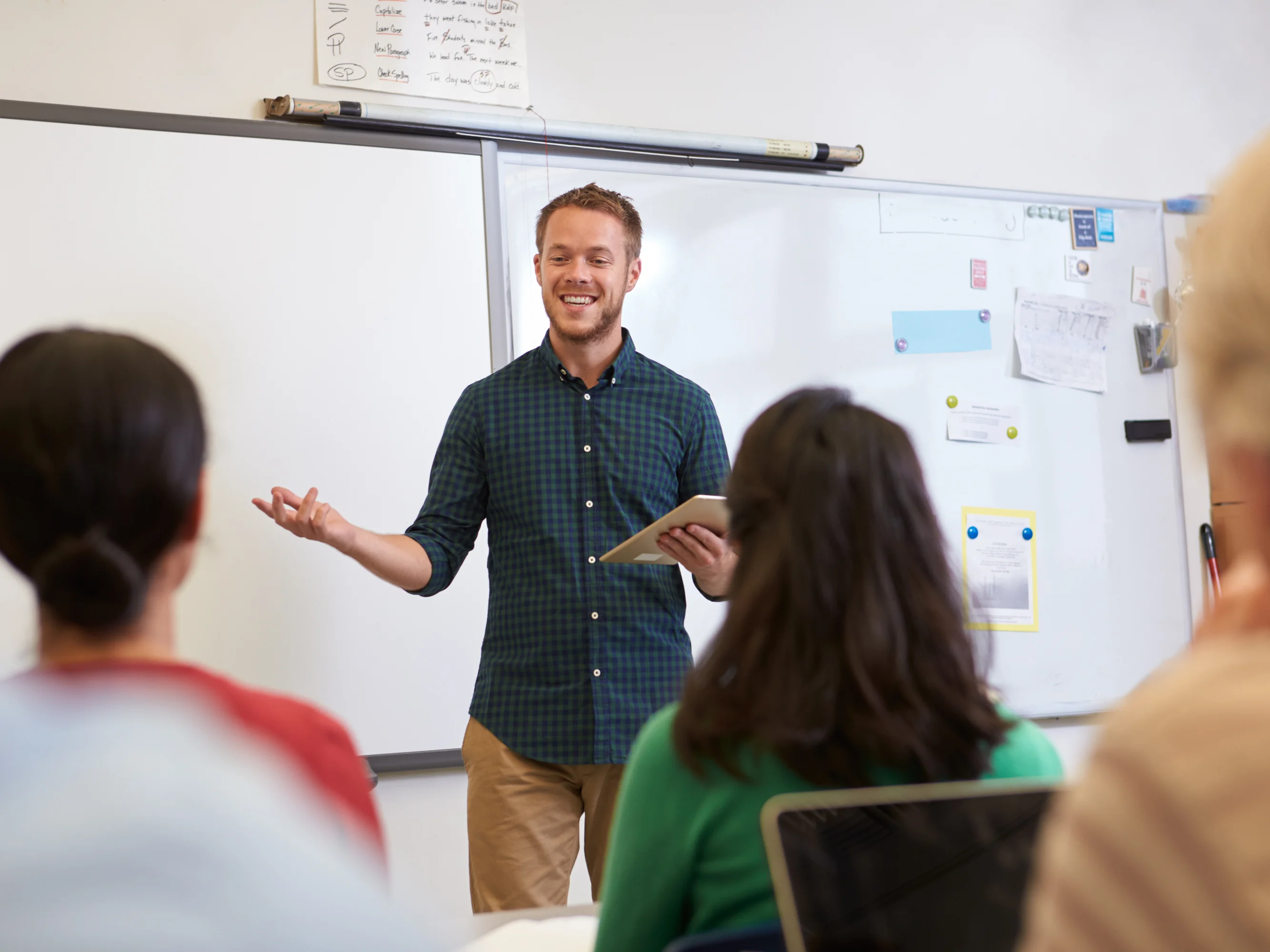 LINC teacher teaching English in a classroom, holding a tablet and smiling. A whiteboard with notes is behind him, while attentive students face him.