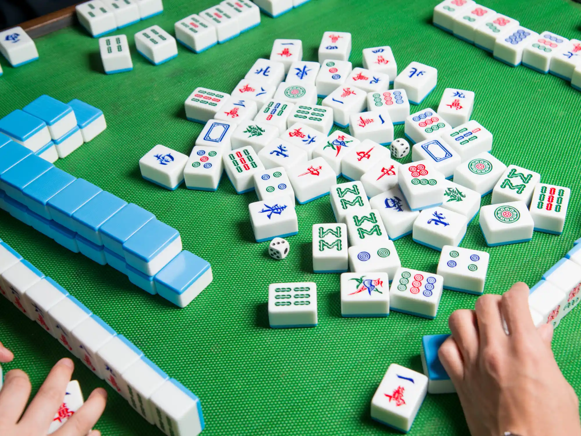 A green table holds scattered Mahjong tiles with colourful symbols, a white die, and hands arranging tiles.