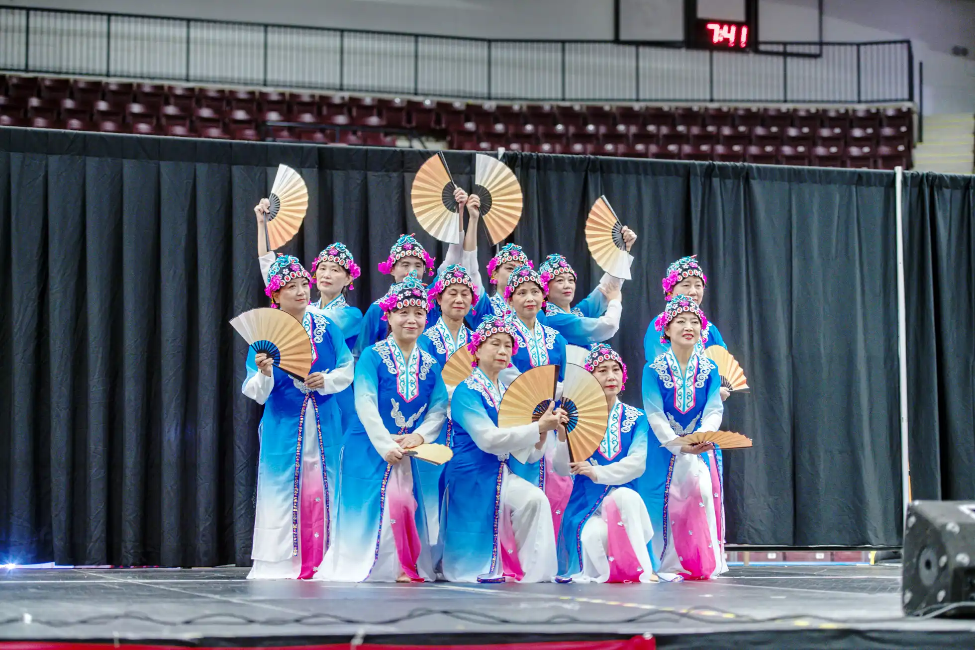 Group of women in blue and pink traditional outfits holding fans arranged in a formation on stage.