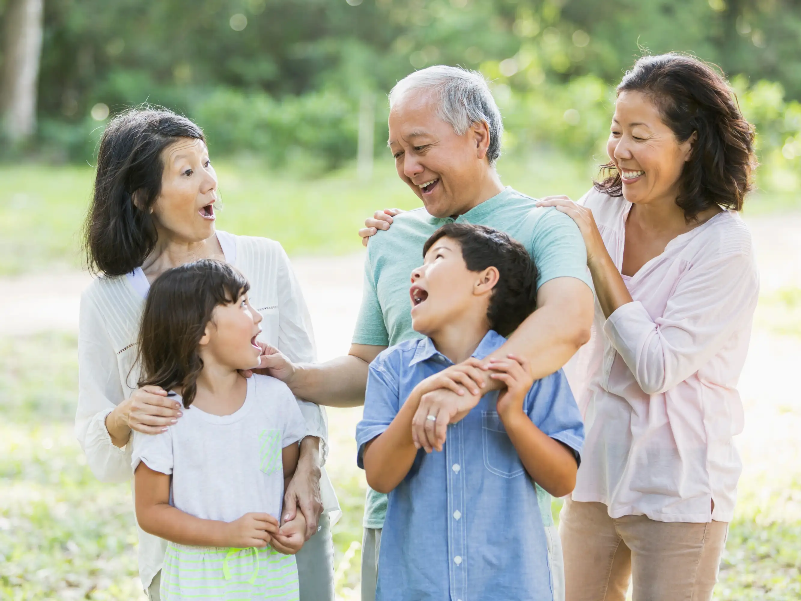 A joyful family of six, including grandparents, parents, and two children, shares a happy moment in a green park, all smiling and laughing together.