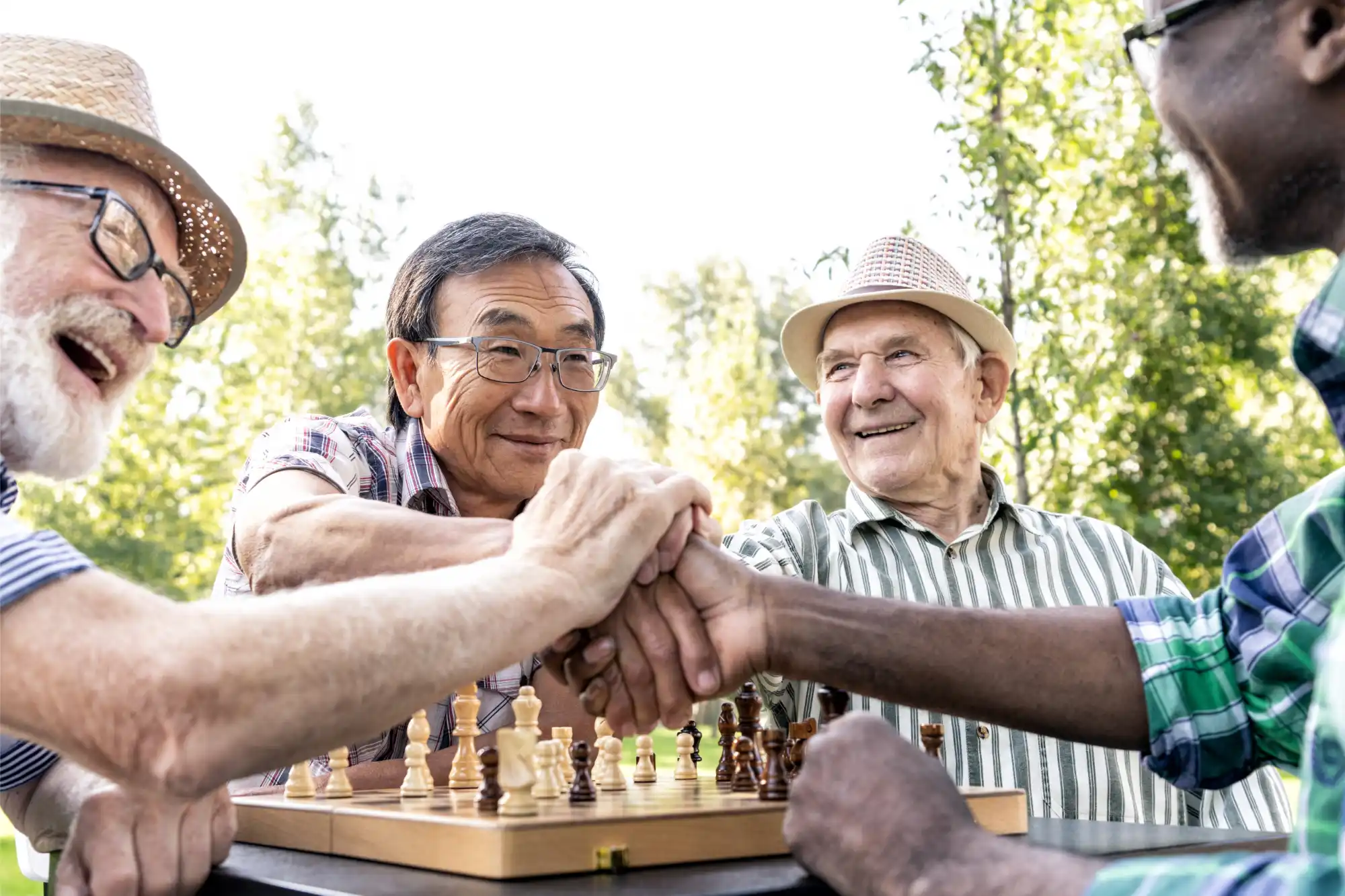 Four elderly men, smiling and joyful, clasp hands over a chessboard in a sunny park. Their hats and plaid shirts convey camaraderie and leisure.