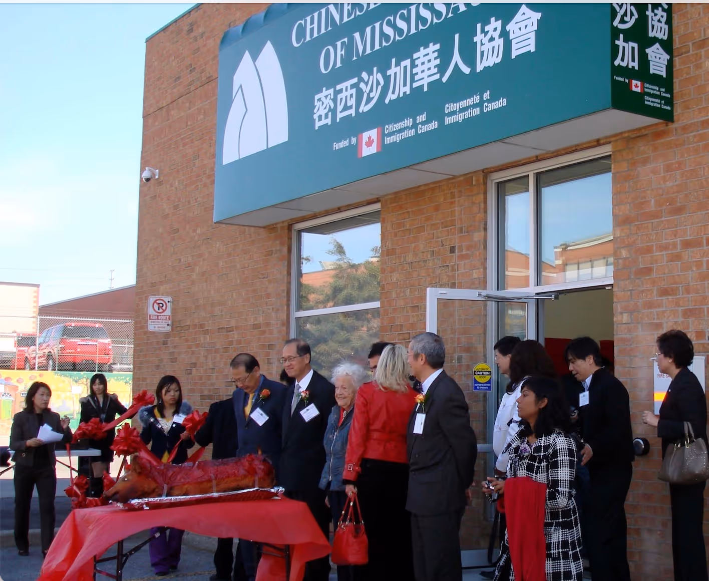 A group gathers outside the Chinese Association of Mississauga building during a ceremony. A roasted pig sits on a red-draped table, symbolizing celebration.