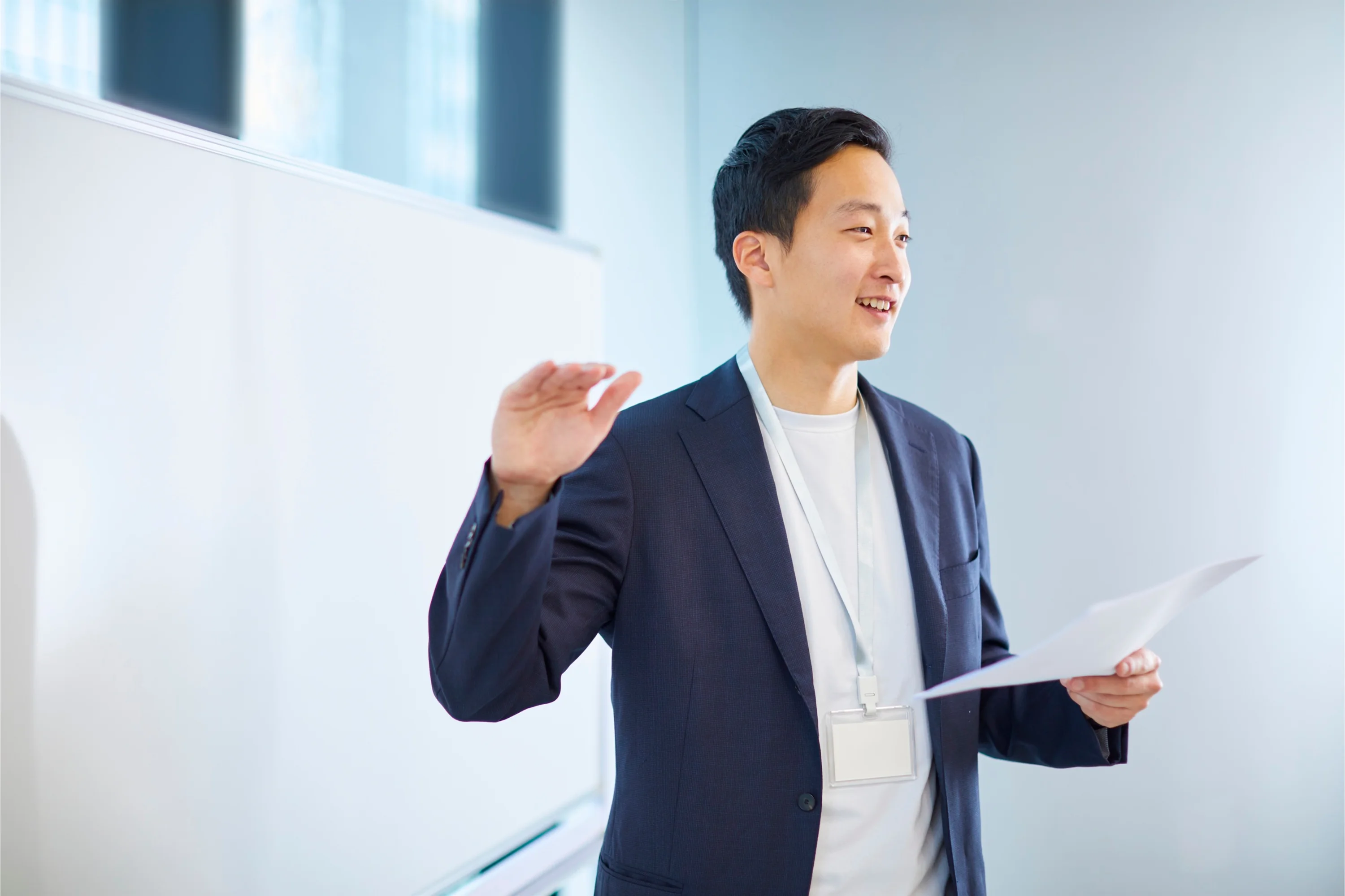 A person in a navy blazer holds a paper, speaking animatedly in a bright room. A whiteboard is visible, suggesting a presentation setting.