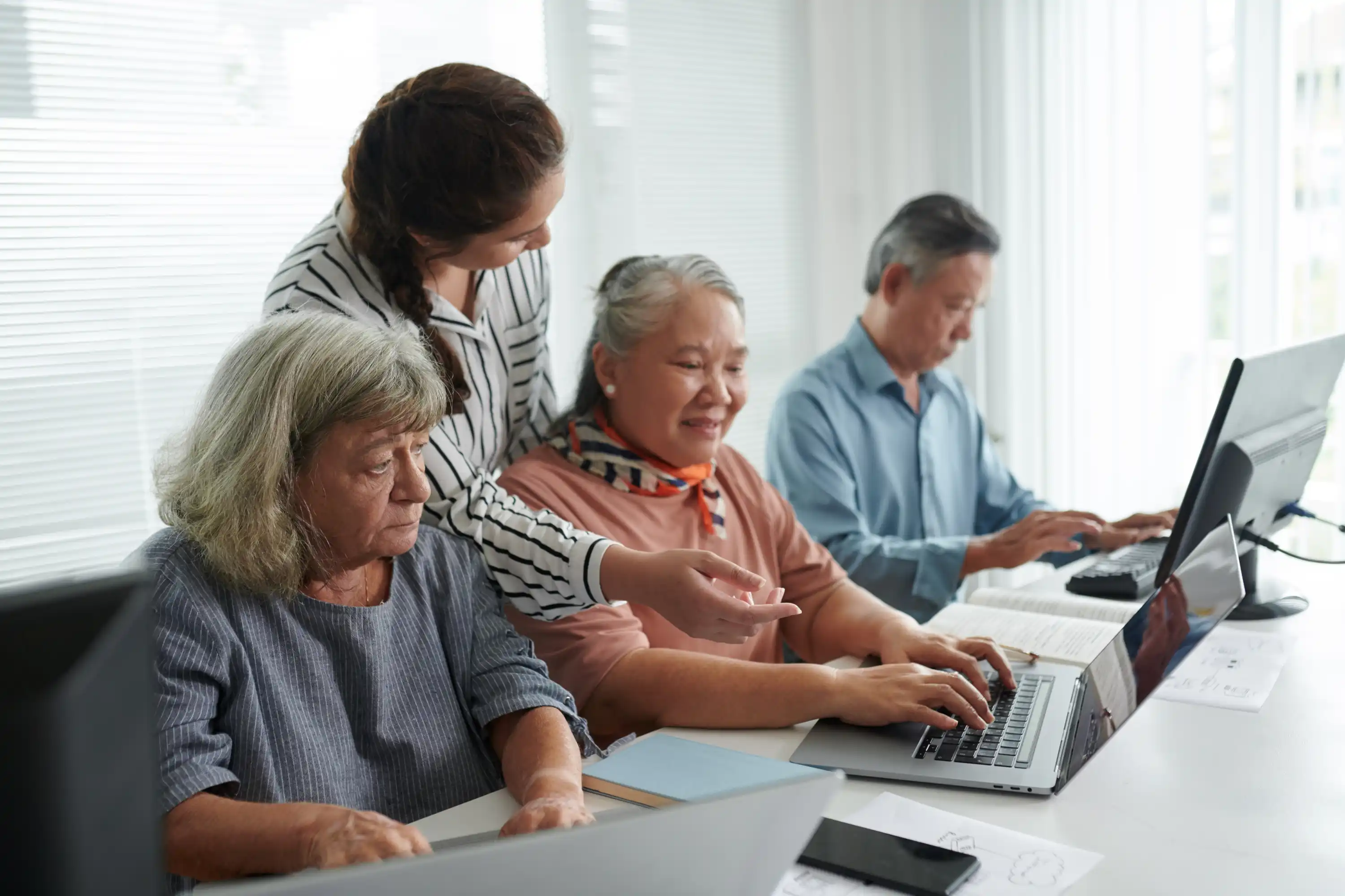 A young woman assists an older woman with a laptop, surrounded by focused elderly learners on computers in a bright, supportive classroom.