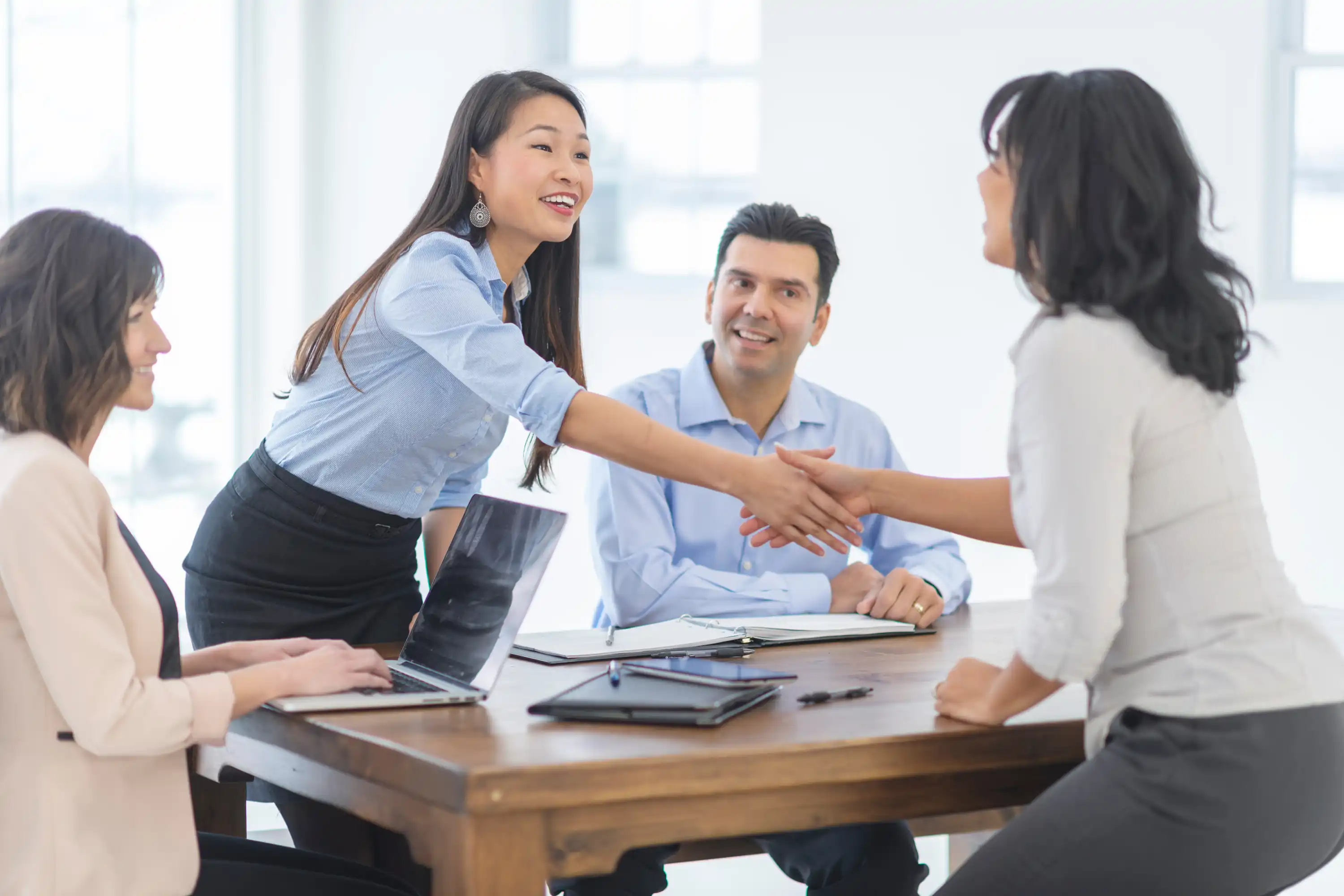 Four professionals in a bright office setting. A woman stands to shake hands with another, while two colleagues sit, smiling. Friendly and collaborative mood.