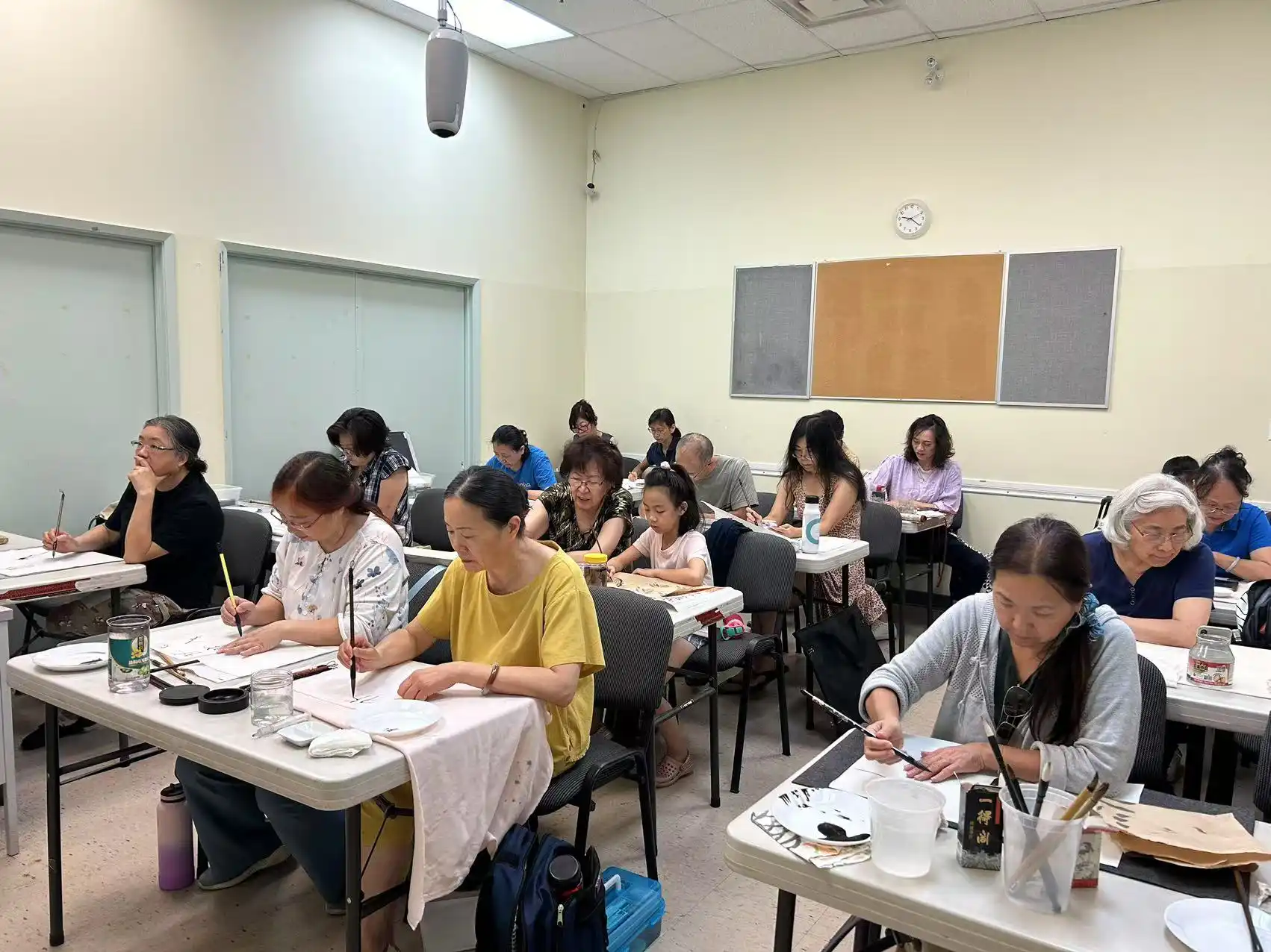 Participants of various ages sit at tables in a classroom setting during a traditional Chinese calligraphy and brush painting class hosted by the Chinese Association of Mississauga. They are actively practicing brush strokes on paper using ink wells, water cups, and specialized brushes.