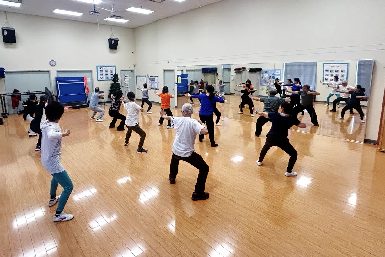 A group of community members, including seniors and adults, practice Tai Chi or martial arts movements in a spacious studio with wood flooring and a large wall mirror. The class, hosted by the Chinese Association of Mississauga, shows participants in a wide stance stretching their arms while following an instructor.