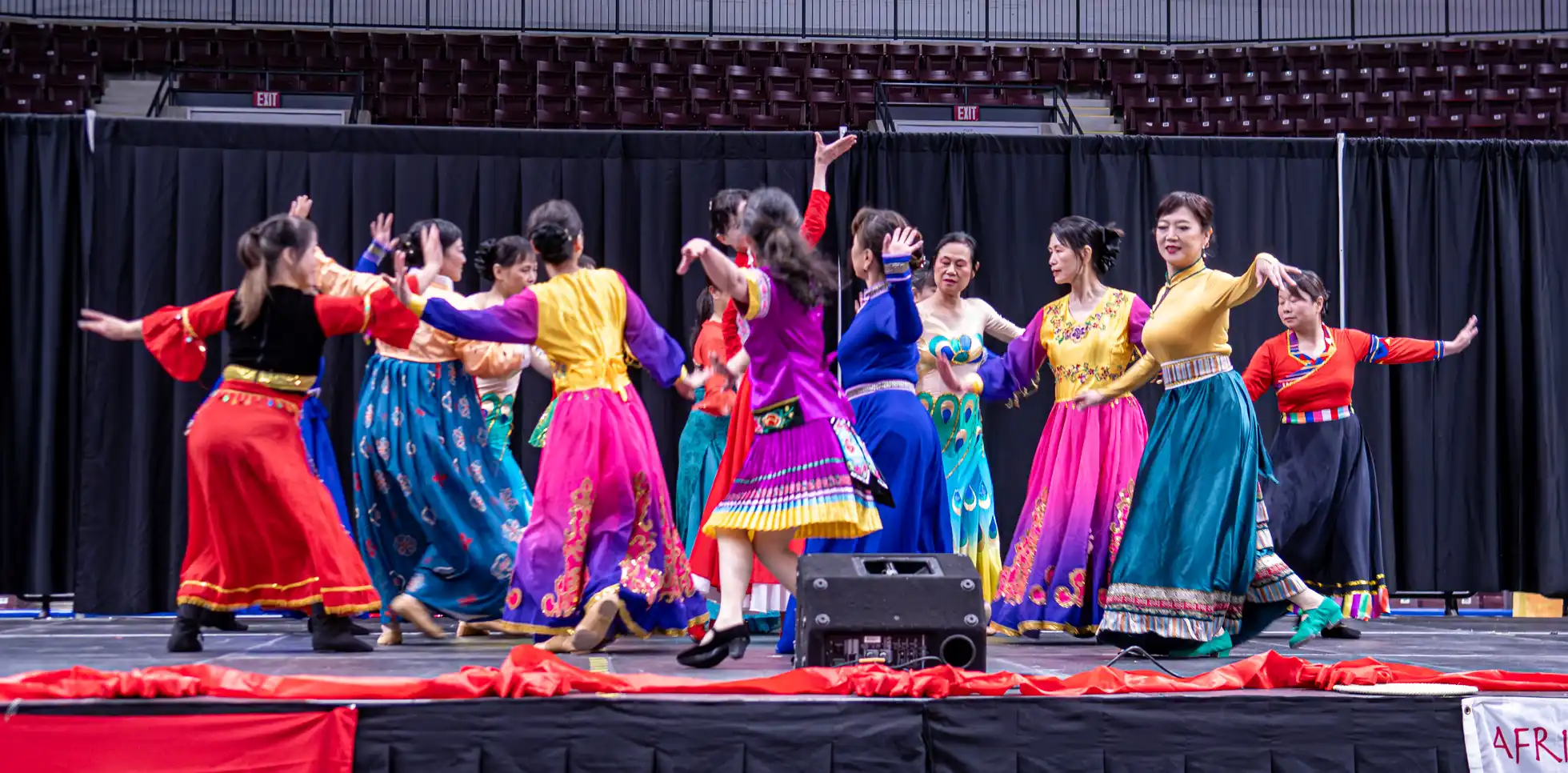 A group of women perform a vibrant Chinese ethnic folk dance on a large stage with a black backdrop. The dancers wear a diverse variety of colorful traditional costumes representing different regional styles, including embroidered skirts, bright silk dresses, and a peacock-patterned gown, hosted by the Chinese Association of Mississauga.