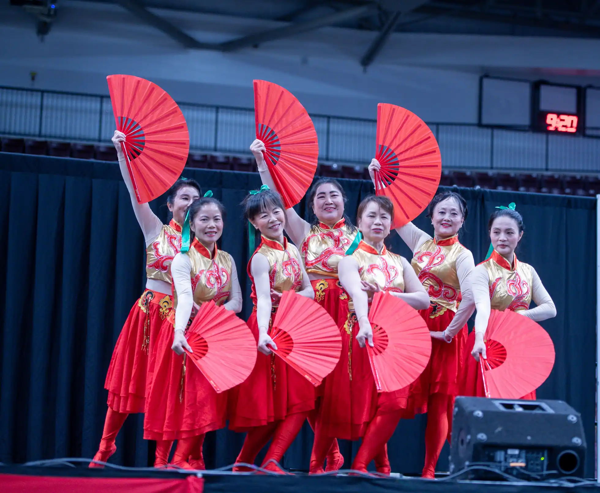 Seven women in red and gold costumes holding red fans posing in a Chinese traditional dance performance.