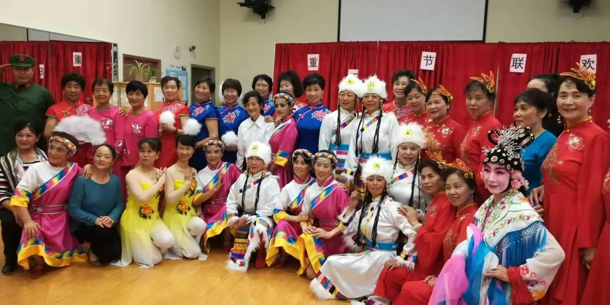 Group of women in colourful traditional Chinese costumes posing indoors against red curtains.