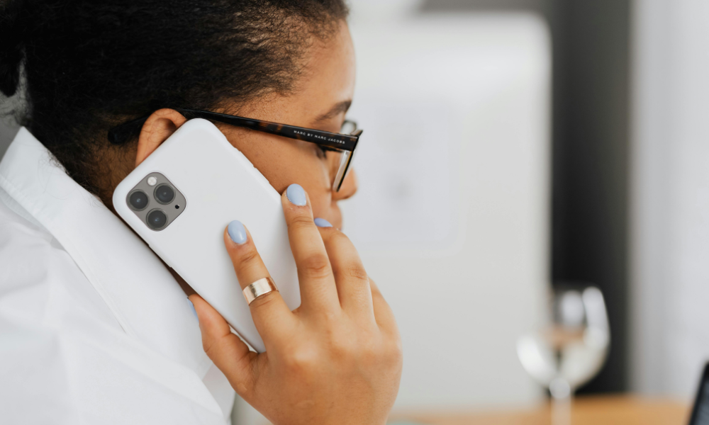 A person wearing glasses and a white shirt is talking on a white smartphone. They have a ring on their finger and light blue nail polish.