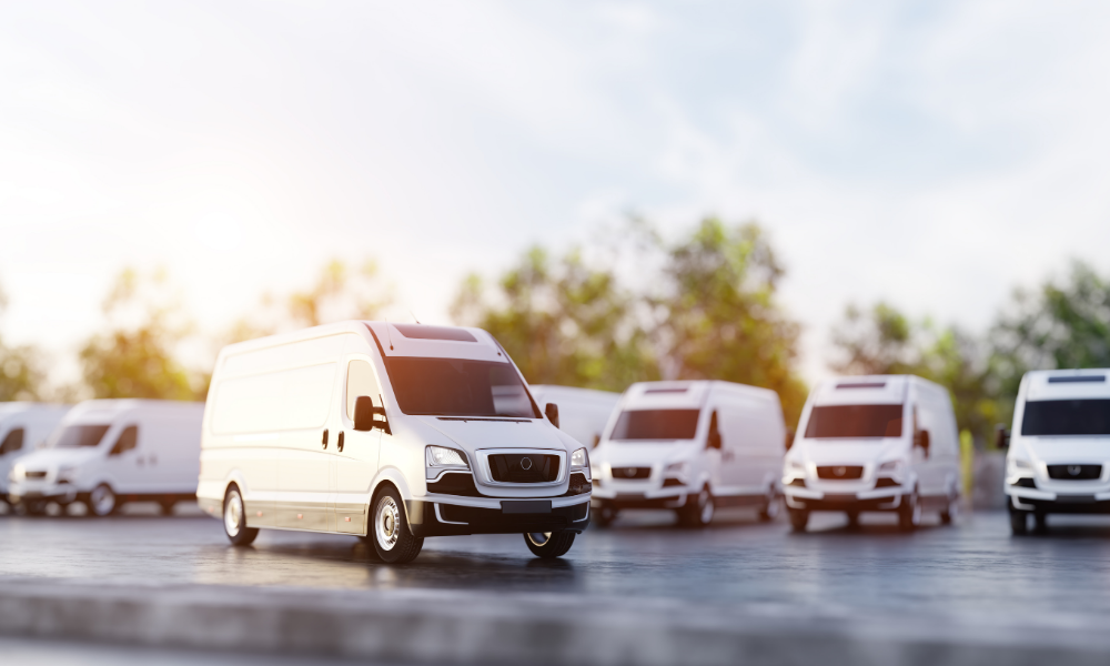 Row of white delivery vans parked on a paved lot, with one front van in sharp focus and others lined up behind it under warm daylight.