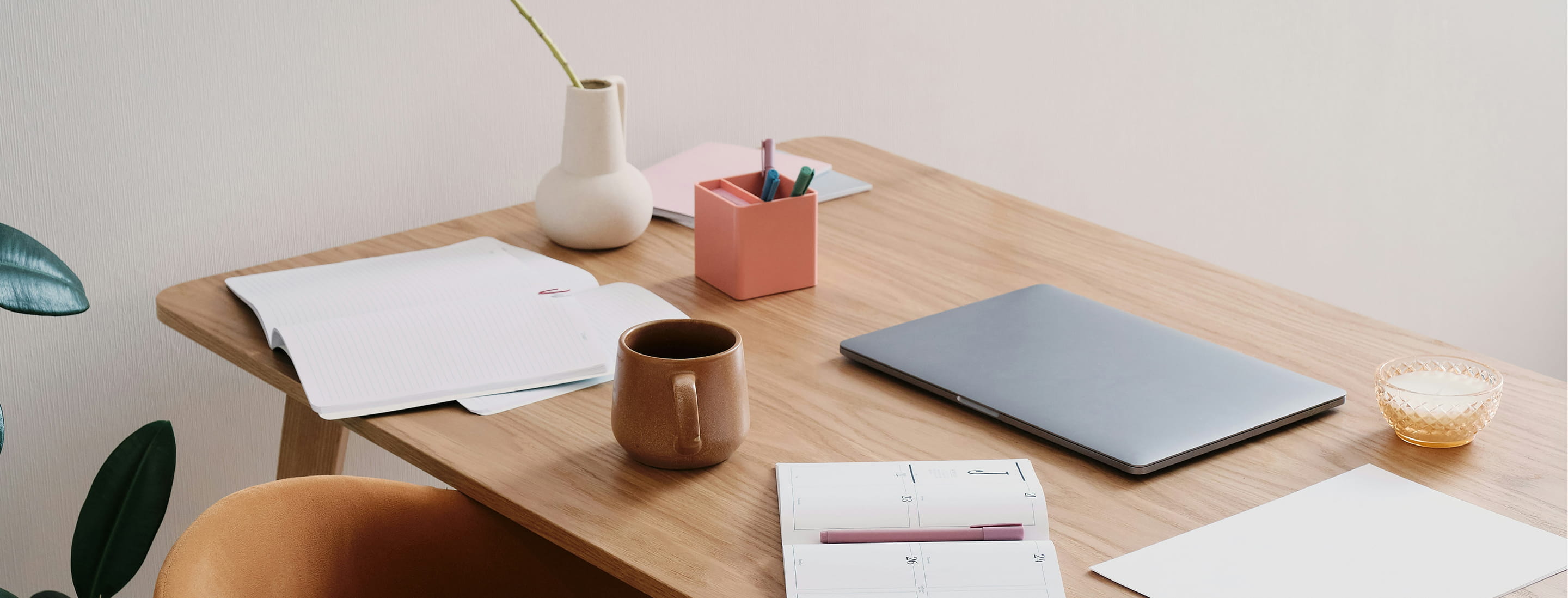 Minimalist wooden desk with a closed laptop, open notebooks, a ceramic mug, pink pen holder, white vase with a single flower stem, and a small glass candle holder.