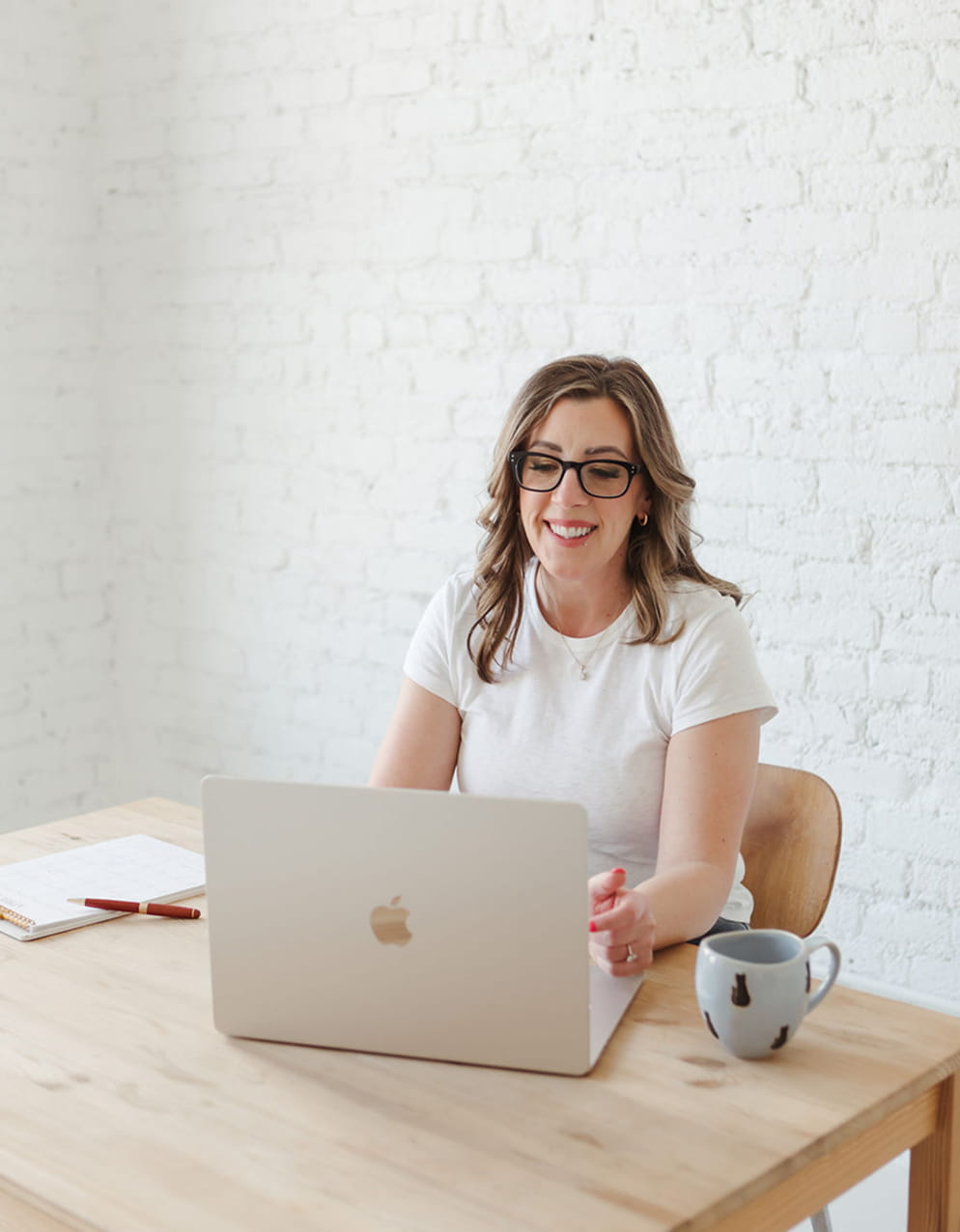 Smiling Sandra Mills with glasses sitting at a wooden table working on a silver Apple laptop with a notebook, pen, and mug nearby.