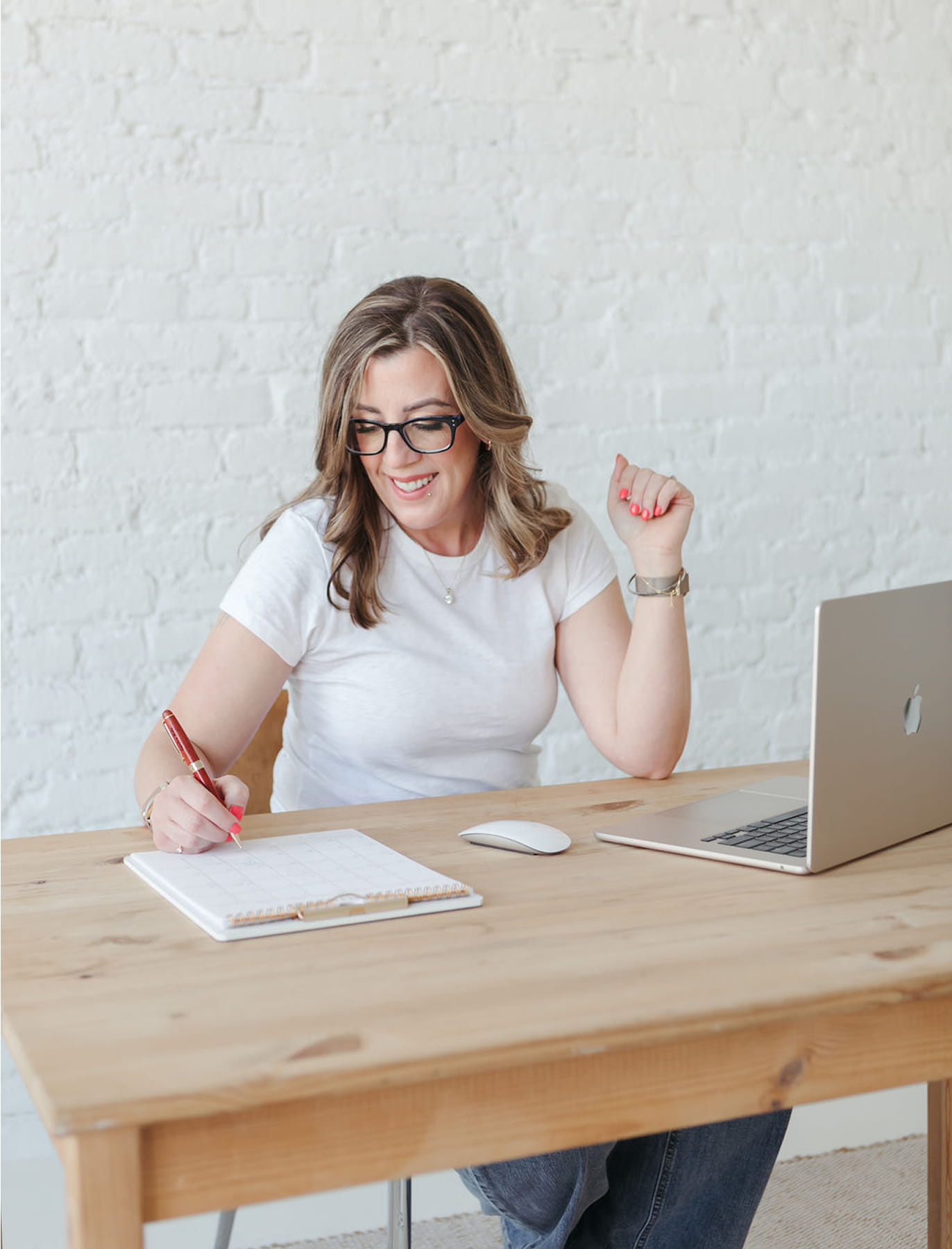 Smiling woman with glasses writing on a calendar notebook at a wooden desk with a laptop and mouse.