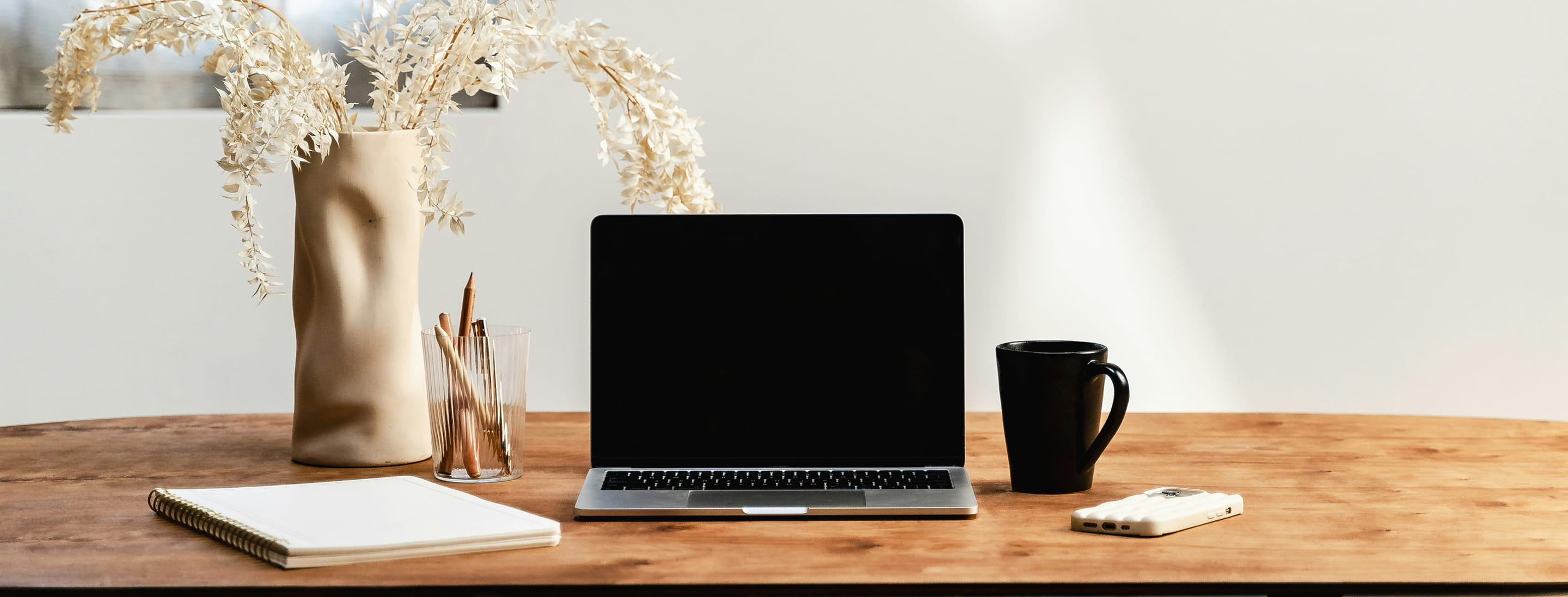 Desk with a closed laptop, a black coffee mug, a white smartphone, a notebook, a glass with pencils, and a vase with dried plants.