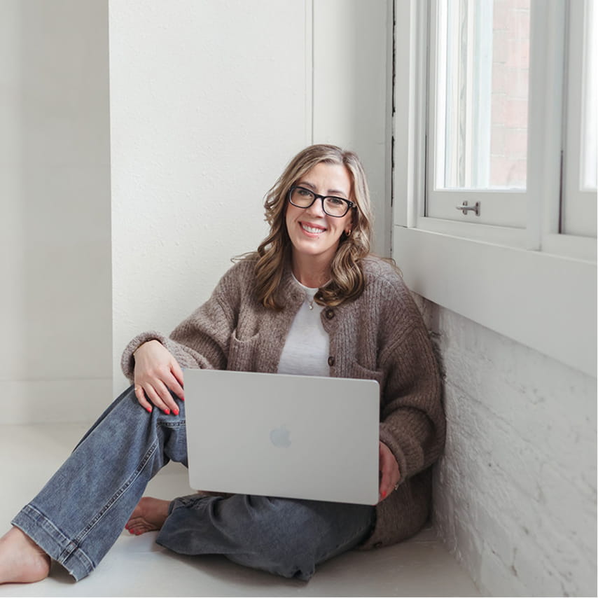 Smiling woman with glasses sitting barefoot on the floor near a window using a laptop.