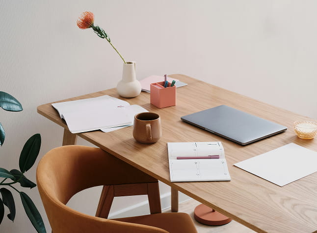 Minimalist wooden desk with a closed laptop, open planner, coffee cup, papers, a pink pen holder, a vase with a single flower, and a brown chair.