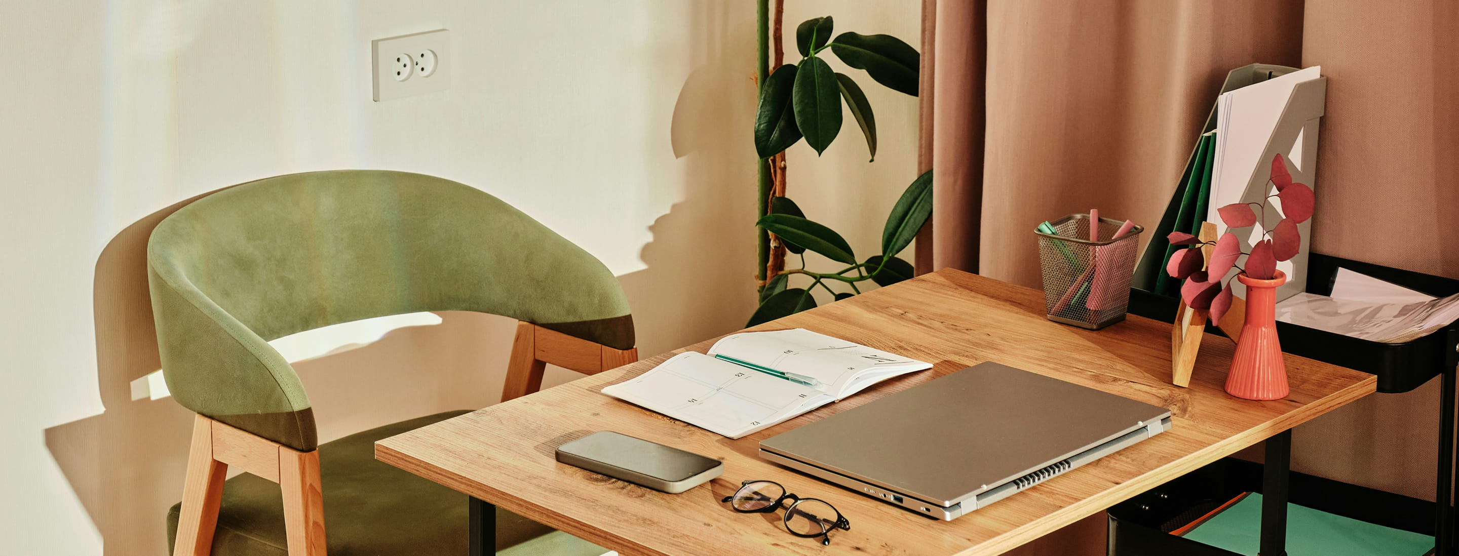 Wooden desk with a closed laptop, open planner with a pen, glasses, a small case, a plant, and stationery holder next to a green cushioned chair.