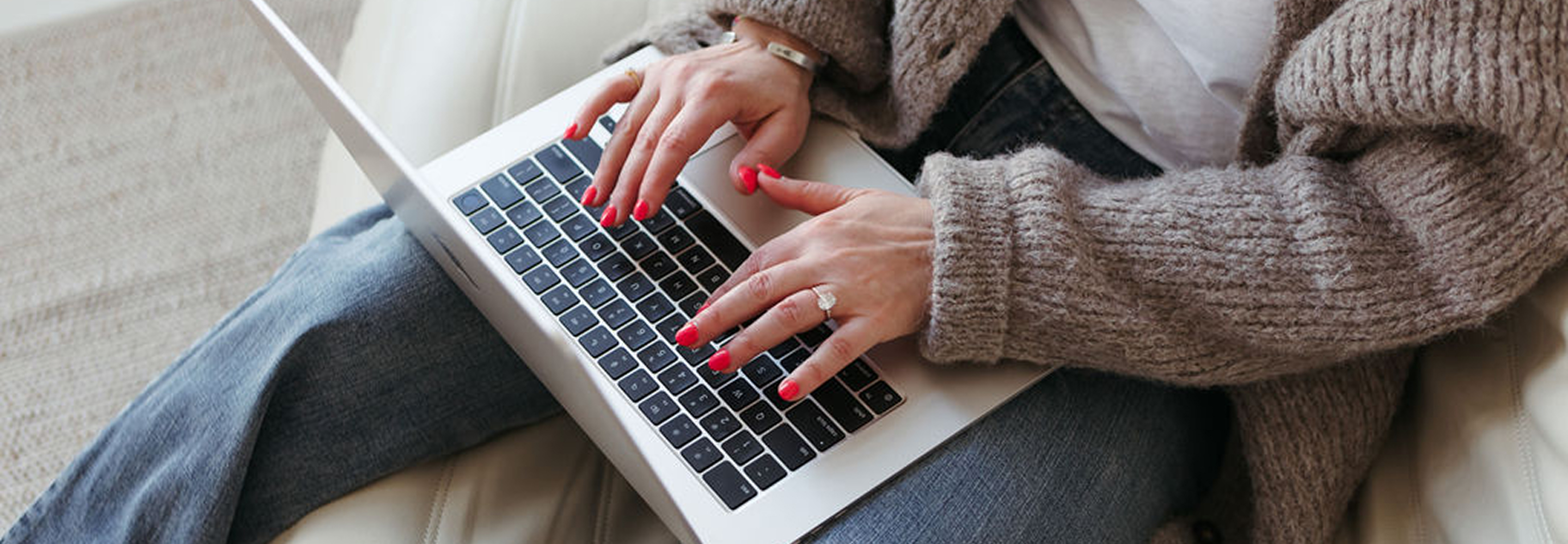 Sandra Mills with red nail polish typing on a laptop keyboard while sitting, wearing a cozy knitted sweater and jeans.
