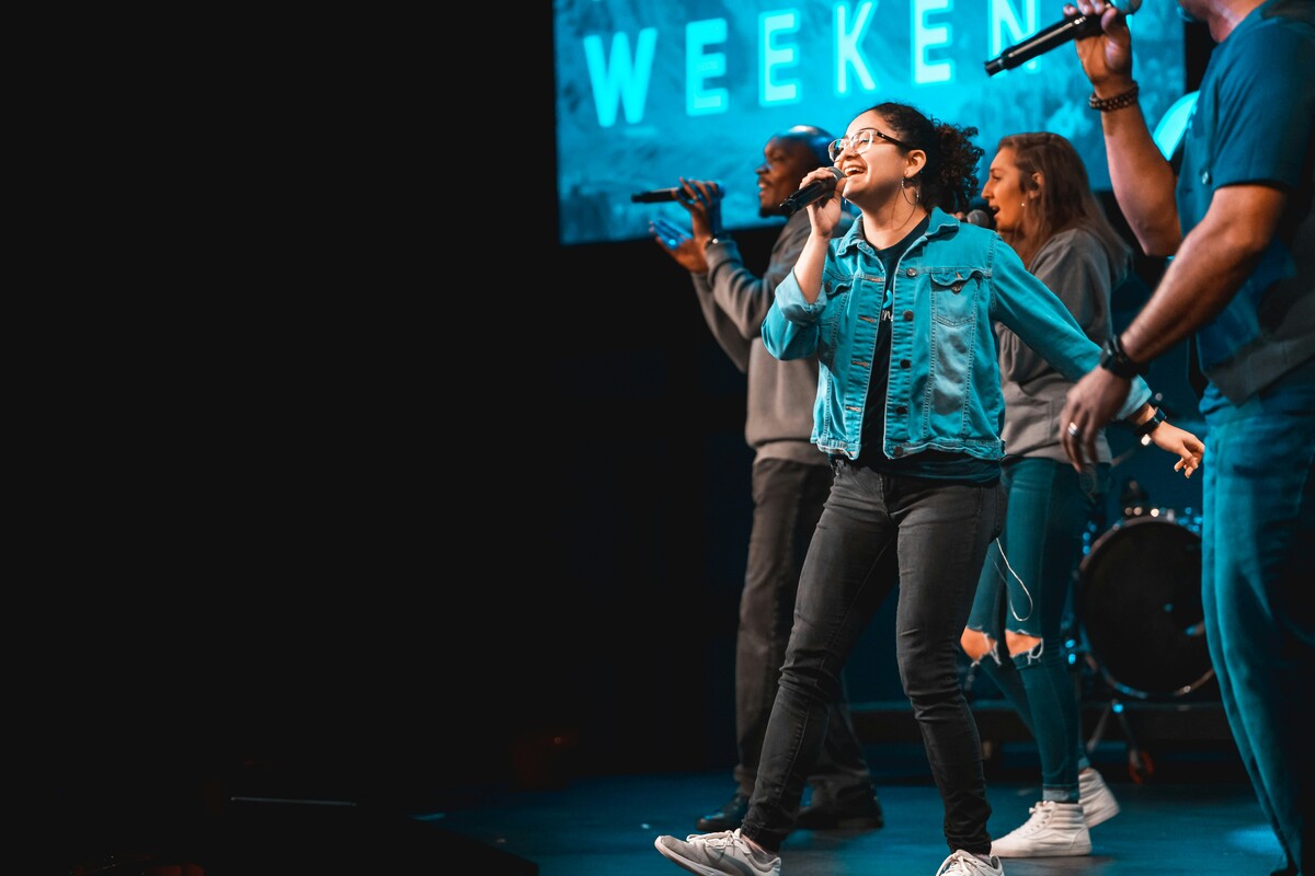 A group of four singers performs on stage with microphones and a 'WEEKEND' sign behind them.