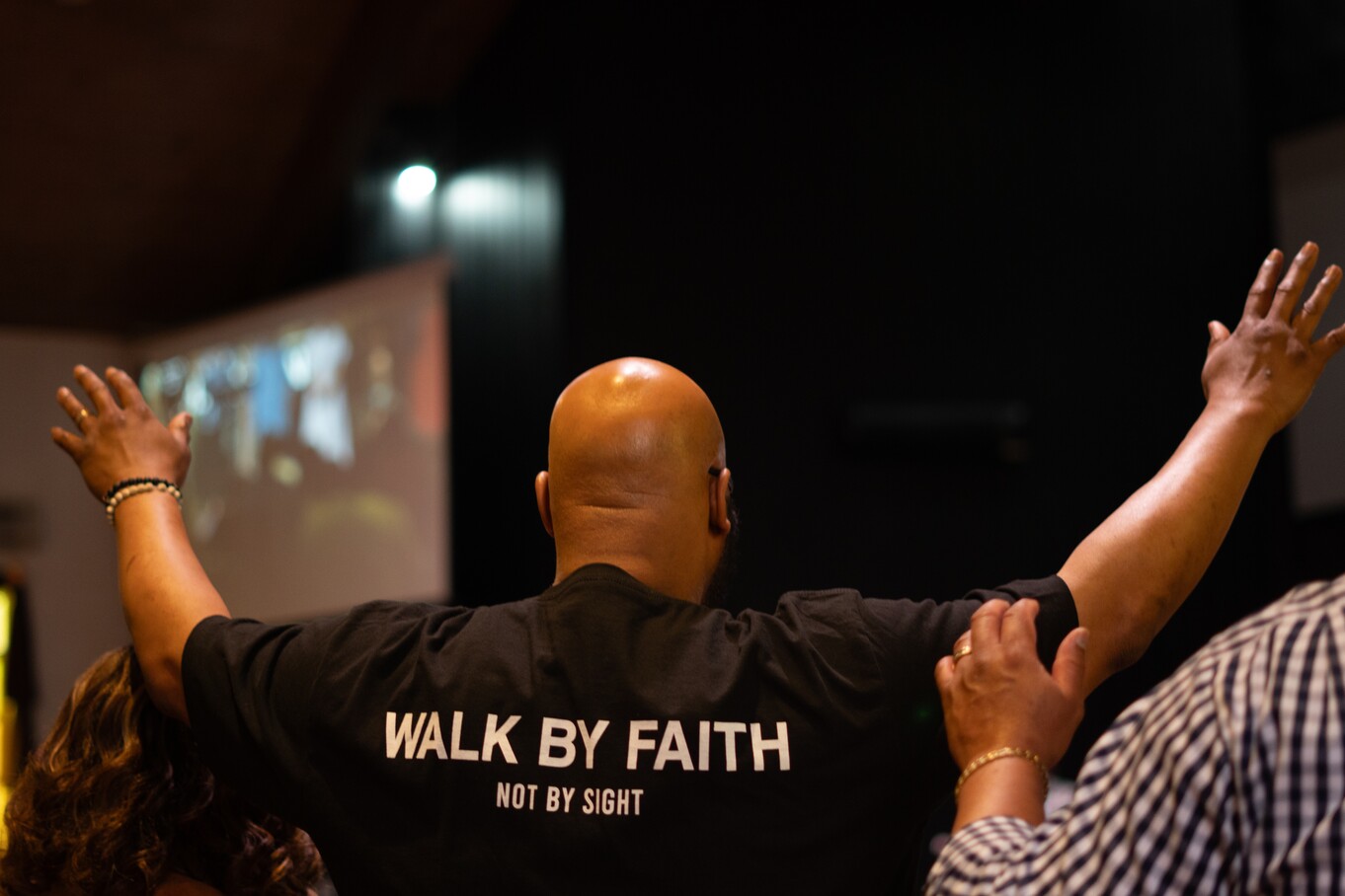 Man raising hands in worship wearing a black shirt that says WALK BY FAITH NOT BY SIGHT.