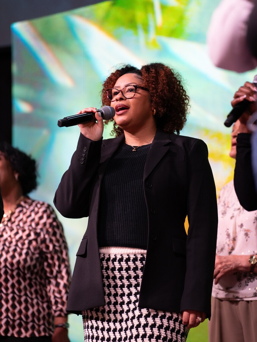 Woman with curly hair and glasses singing into a microphone on stage with colorful background.