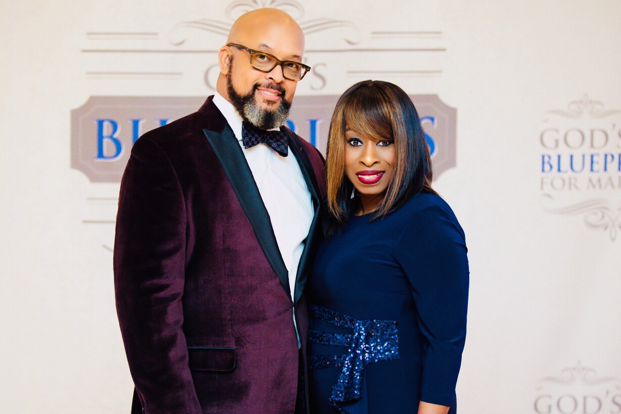 Man in burgundy velvet suit and bow tie stands next to woman in navy blue dress with sequined belt.