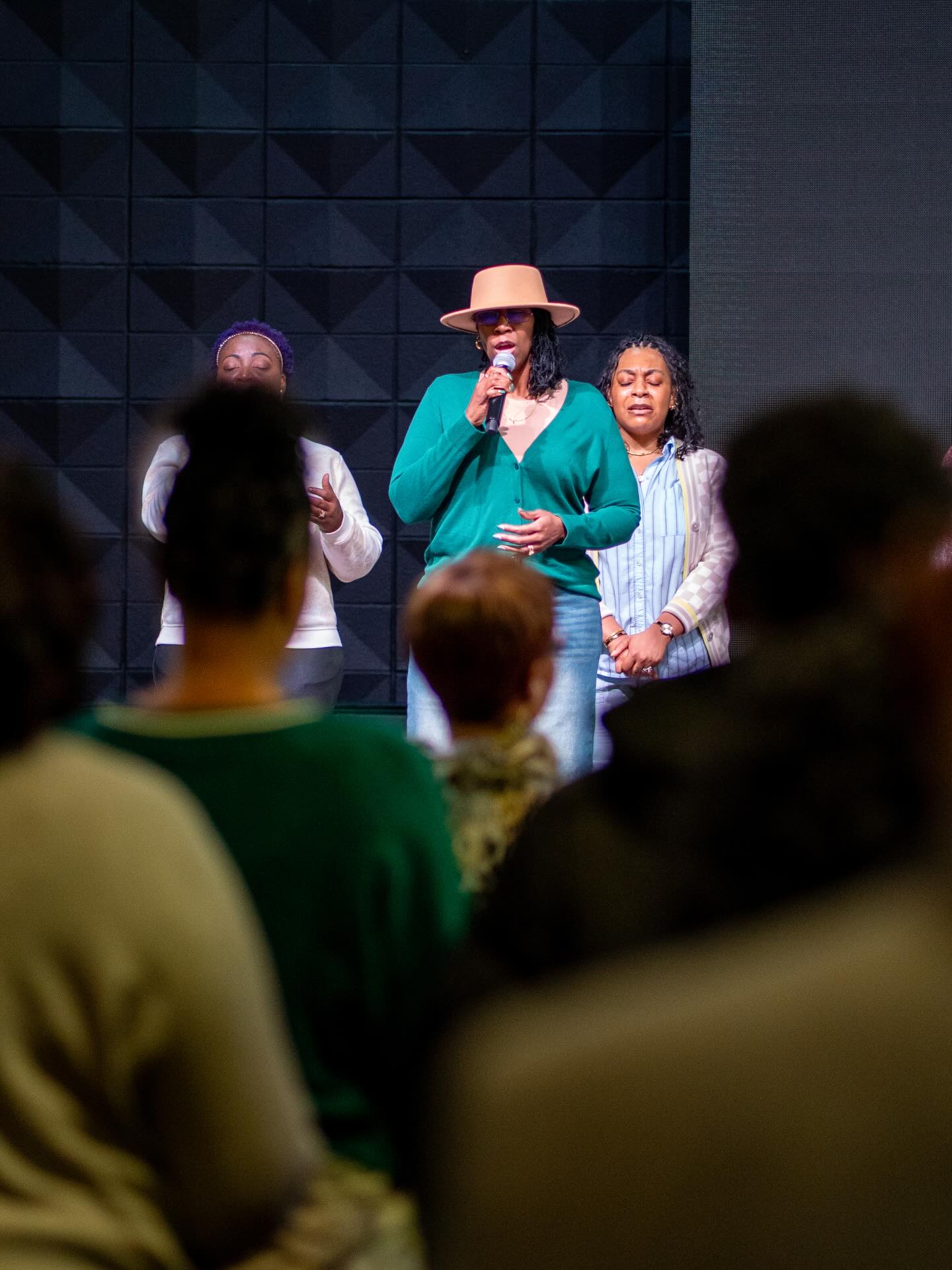 Woman in hat singing into microphone on stage with two women behind her and audience in front.