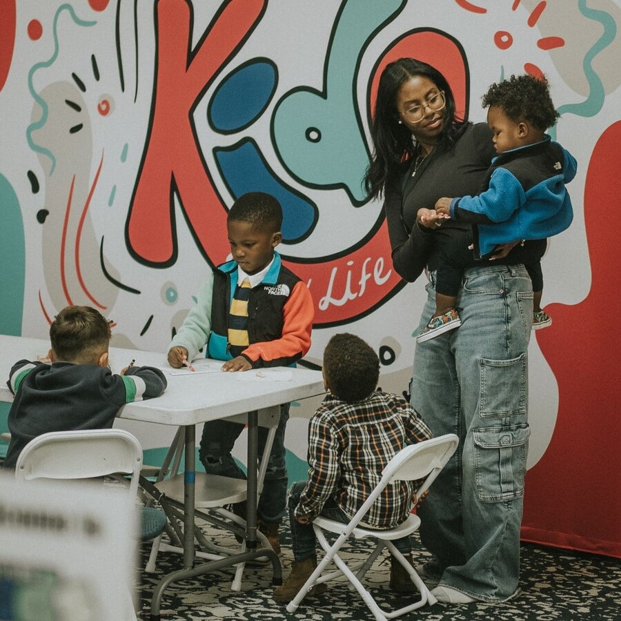 Woman holding toddler interacts with three young boys seated and standing around a table in front of colorful wall art.