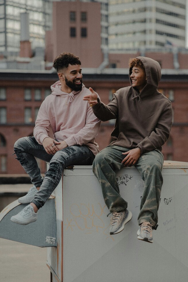 Two young men in hoodies sitting on a rooftop box, smiling and talking with an urban background.