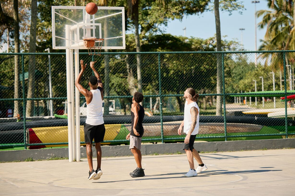 Three men playing basketball outdoors with one jumping to shoot the ball towards the hoop.