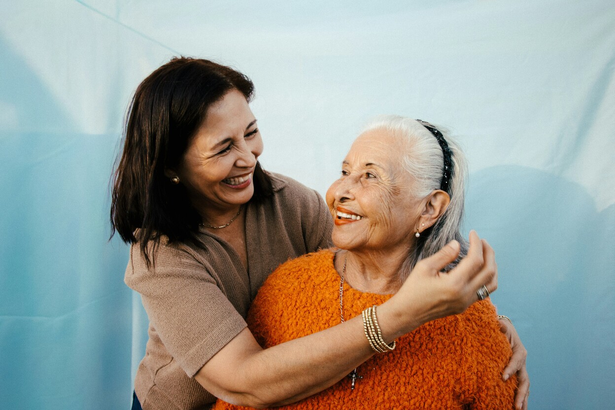 Smiling middle-aged woman embracing and looking at happy elderly woman in an orange sweater.