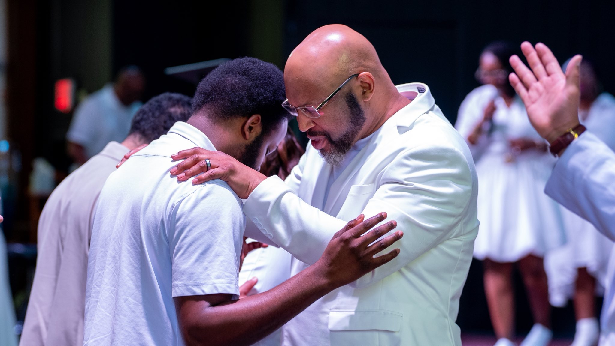 Man in white jacket comforting and praying with another man in a white shirt in a church setting.