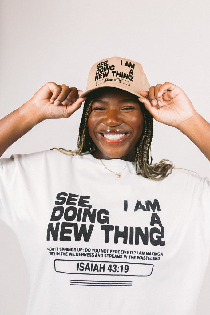 Smiling woman with braided hair wearing a hat and T-shirt with Isaiah 43:19 Bible verse text.