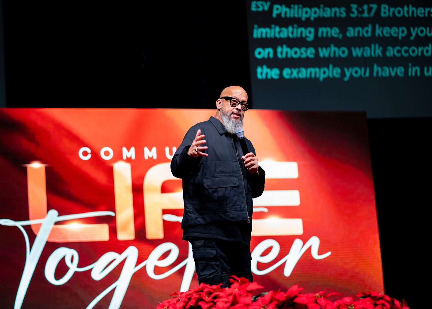 Man with glasses and gray beard speaking on stage with a microphone and red flowers in front.