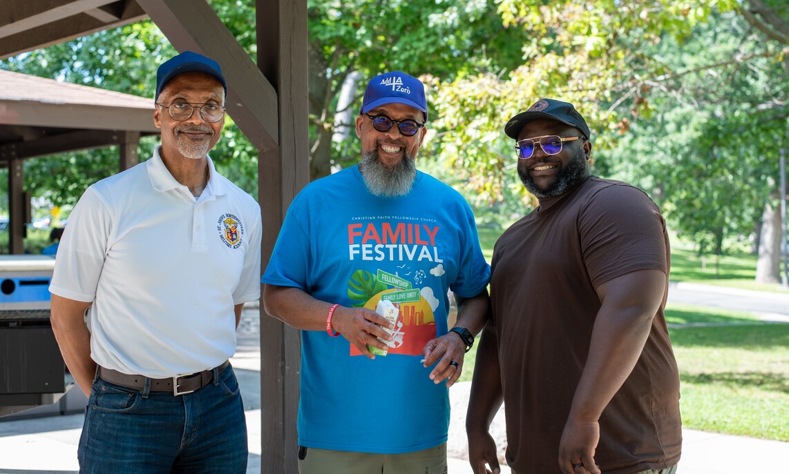 Three men smiling outdoors under a wooden pavilion with trees in the background.
