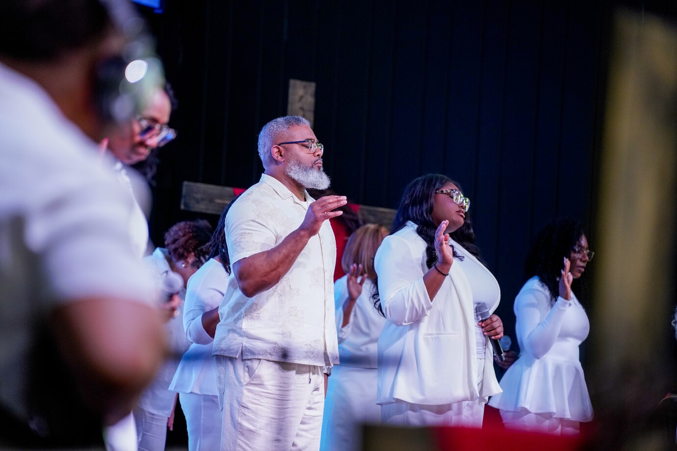Diverse group in white clothes singing and worshiping with hands raised in church setting.