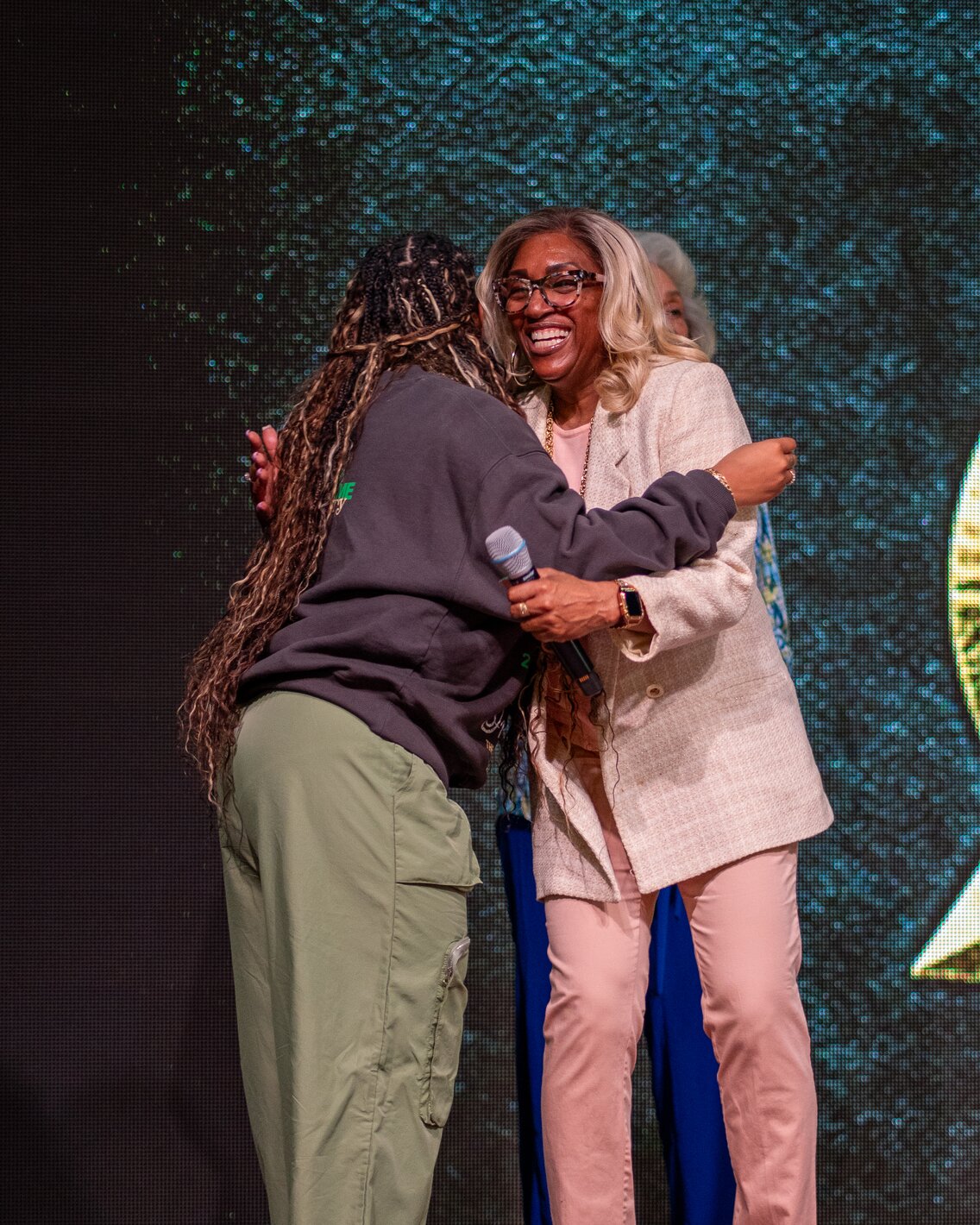 Two women warmly hugging on stage, one holding a microphone, with a digital screen background.