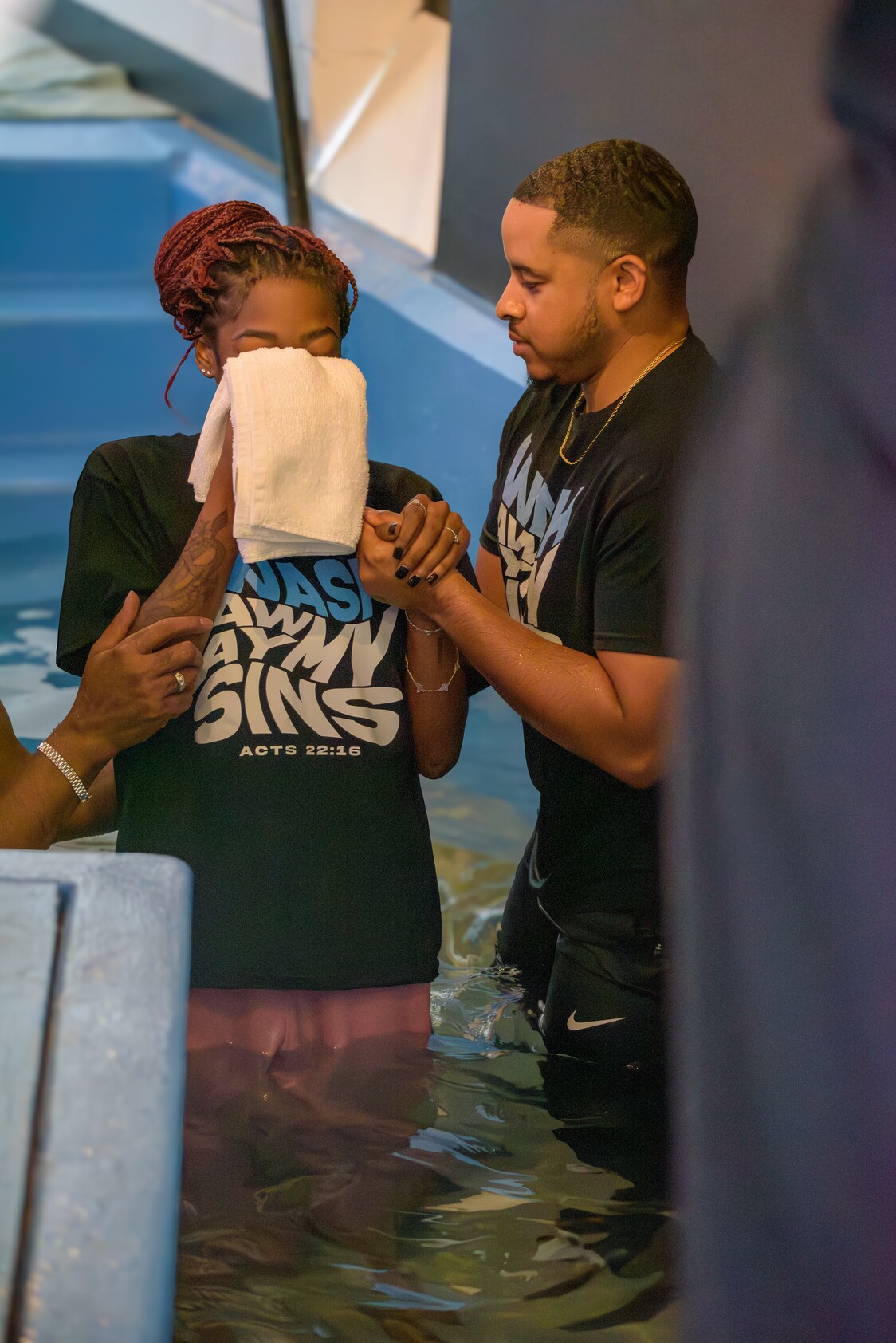 A woman is being baptized in a pool, holding a towel to her face, supported by two people.