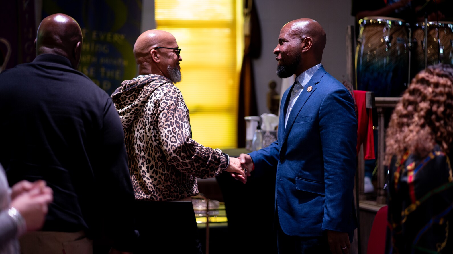 Two men shaking hands indoors, one in a blue suit and the other in a leopard print hoodie.