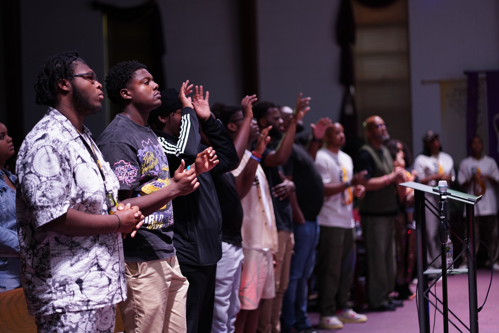 Group of people standing indoors with hands raised, appearing focused or in prayer near a podium.