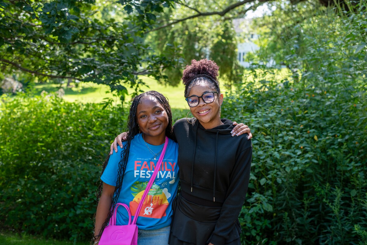 Two women stand outside with arms around each other, smiling in front of green foliage.