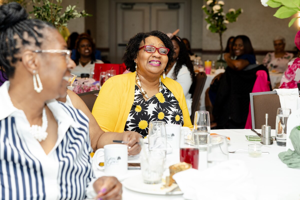 Smiling woman in yellow cardigan and flower dress sitting at a banquet table with others.