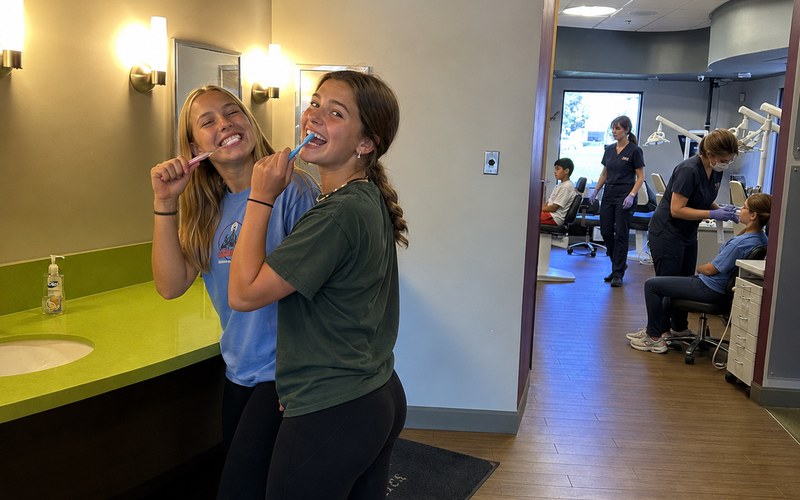 Two teens brushing their teeth in the hygiene area at Kendrick Orthodontics while patients and assistants are in the treatment area behind them.