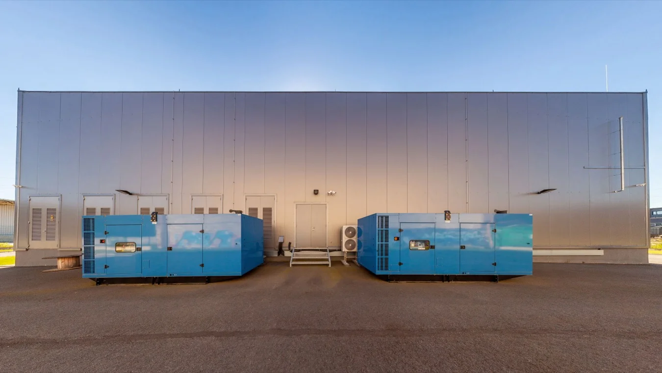 Two blue generator units outside a metal-clad industrial building at dusk