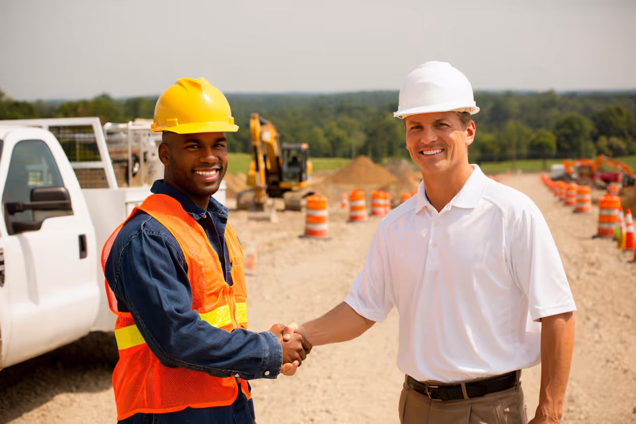 Two construction workers wearing safety helmets and shaking hands at a construction site with traffic barrels and equipment in the background.